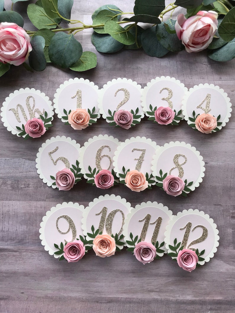 May include: Twelve white scalloped circles with pink and peach paper flowers and green leaves. Each circle has a gold glitter number from one to twelve.