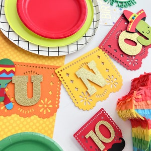 May include: A colorful fiesta party banner with the letters "UNO" and the number "10" in gold glitter. The banner is decorated with maracas, a cactus, and a pinata.