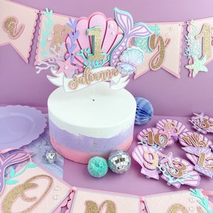 Peut inclure: Un g&acirc;teau rose et violet avec un d&eacute;cor sur le th&egrave;me de la sir&egrave;ne. Le d&eacute;cor comprend une coquille Saint-Jacques, une queue de sir&egrave;ne et le chiffre un. Le g&acirc;teau est d&eacute;cor&eacute; de paillettes dor&eacute;es et le nom "Julianna" est &eacute;crit sur une banni&egrave;re. Il y a &eacute;galement plusieurs d&eacute;corations sur le th&egrave;me de la sir&egrave;ne autour du g&acirc;teau, notamment des coquillages, des &eacute;toiles de mer et une banni&egrave;re qui dit "Un".