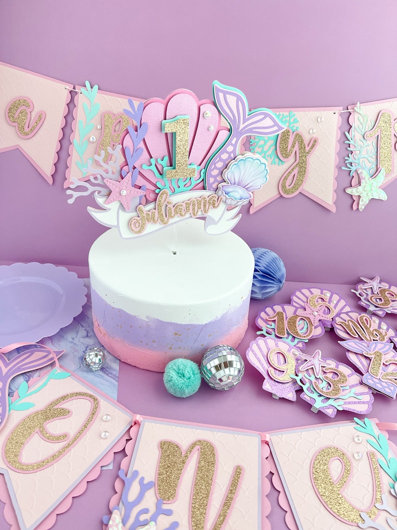 Peut inclure: Un g&acirc;teau rose et violet avec un d&eacute;cor sur le th&egrave;me de la sir&egrave;ne. Le d&eacute;cor comprend une coquille Saint-Jacques, une queue de sir&egrave;ne et le chiffre un. Le g&acirc;teau est d&eacute;cor&eacute; de paillettes dor&eacute;es et le nom "Julianna" est &eacute;crit sur une banni&egrave;re. Il y a &eacute;galement plusieurs d&eacute;corations sur le th&egrave;me de la sir&egrave;ne autour du g&acirc;teau, notamment des coquillages, des &eacute;toiles de mer et une banni&egrave;re qui dit "Un".