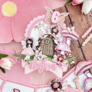 May include: A pink and gold fairy themed cake topper with a mushroom, a door, a bunny, and a banner that says "Meghan". The topper also includes a pink butterfly with a gold glitter accent and the number "6" in pink.