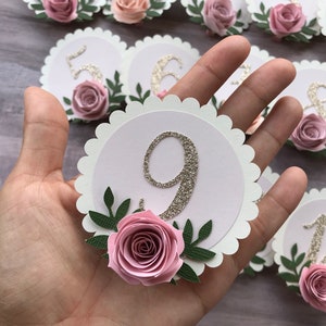 May include: Nine white scalloped circle table number place cards with gold glitter numbers and pink paper roses with green leaves.