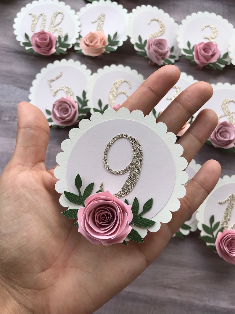 May include: Nine white scalloped circle table number place cards with gold glitter numbers and pink paper roses with green leaves.