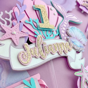 Peut inclure: Un topper de g&acirc;teau sur le th&egrave;me de la sir&egrave;ne en violet, rose et bleu avec un num&eacute;ro un en paillettes dor&eacute;es, le nom "Julianna" en paillettes dor&eacute;es et diverses d&eacute;corations sur le th&egrave;me de la sir&egrave;ne.