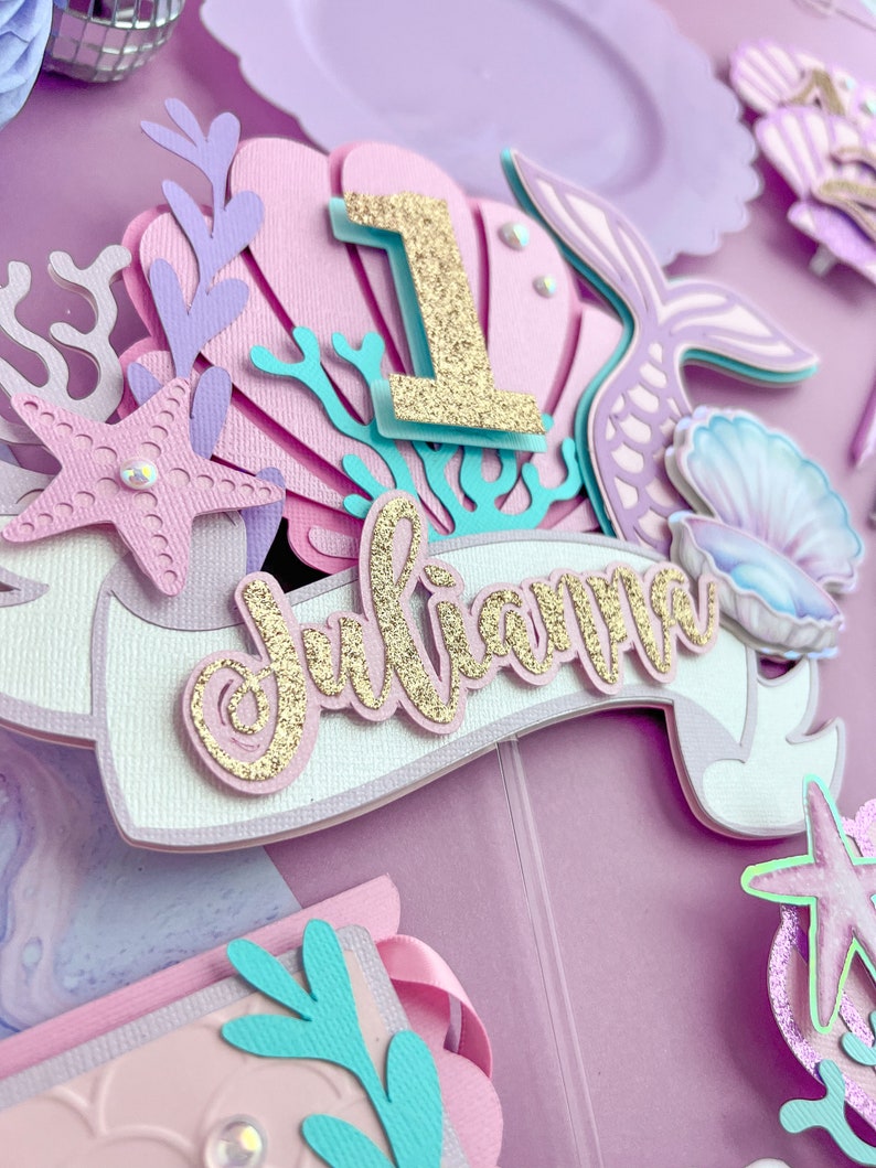Peut inclure: Un topper de g&acirc;teau sur le th&egrave;me de la sir&egrave;ne en violet, rose et bleu avec un num&eacute;ro un en paillettes dor&eacute;es, le nom "Julianna" en paillettes dor&eacute;es et diverses d&eacute;corations sur le th&egrave;me de la sir&egrave;ne.