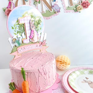 May include: A pink and white cake topper with a bunny rabbit, a number one, and a banner that says "Gabriella". The cake topper is on a pink cake with a pink and white scalloped edge.