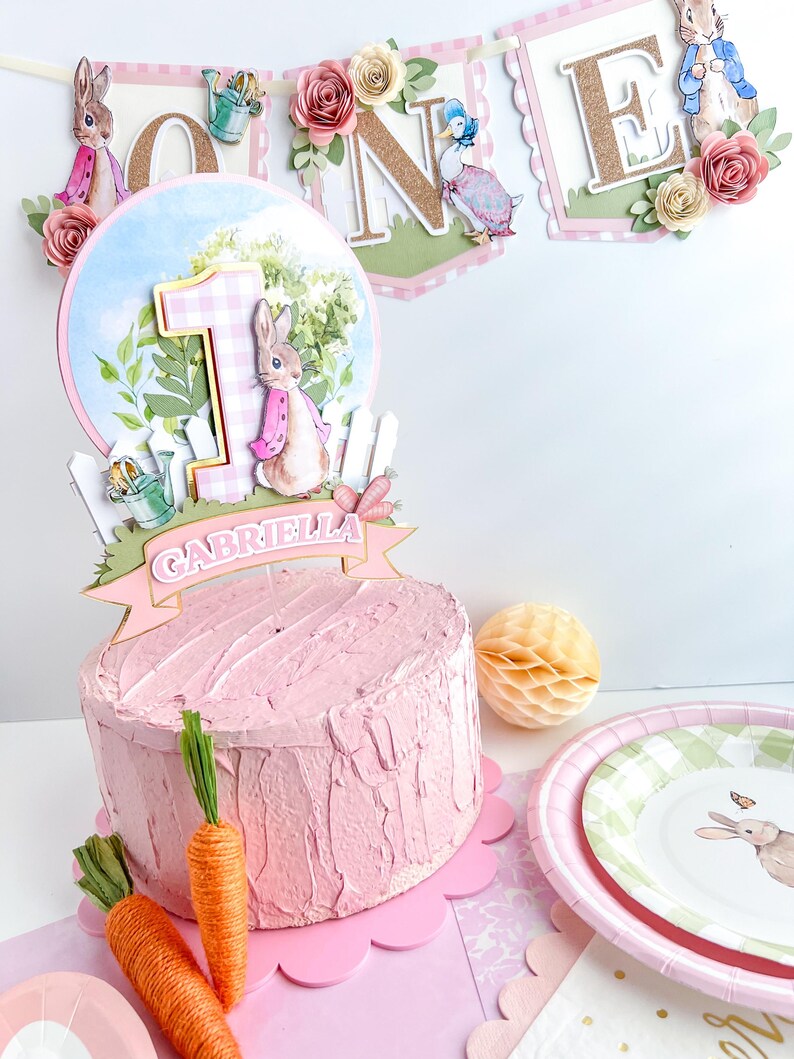 May include: A pink and white cake topper with a bunny rabbit, a number one, and a banner that says "Gabriella". The cake topper is on a pink cake with a pink and white scalloped edge.
