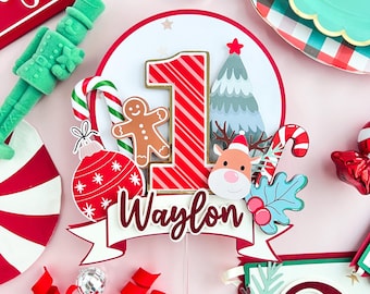 May include: A red, white, and gold Christmas cake topper with a red and white striped number one, a gingerbread man, a candy cane, a Christmas tree, a reindeer, and a holly sprig. The topper also includes a white banner with the name "Waylon" in red.