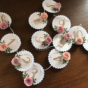 May include: A pink and gold floral garland with numbers 1 through 11. The garland is made of paper circles with scalloped edges and features pink paper flowers and green leaves. The numbers are gold glitter.