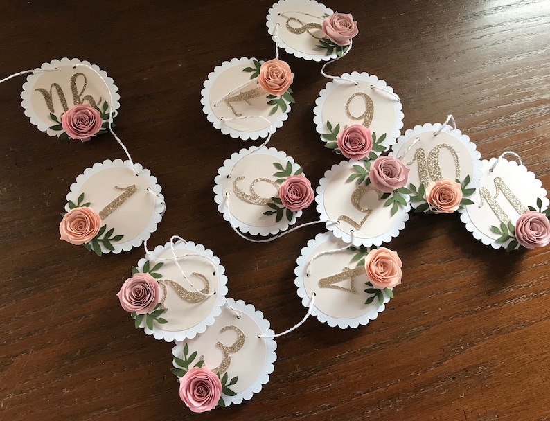 May include: A pink and gold floral garland with numbers 1 through 11. The garland is made of paper circles with scalloped edges and features pink paper flowers and green leaves. The numbers are gold glitter.