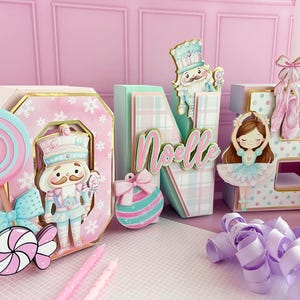 May include: A festive holiday-themed display featuring the word "Noelle" in decorative letters. The letters are adorned with nutcrackers, a ballerina, and a candy theme in pastel pink, blue, and white. A large lollipop and candy accents complete the scene.