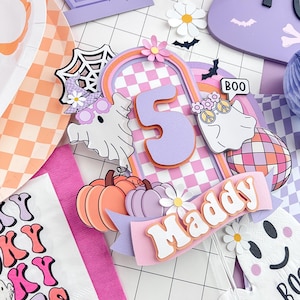 May include: A layered cake topper featuring the number 5 in lavender, a checkered arch, two ghosts with accessories, a disco ball, pumpkins, and a banner with the name Maddy. The design includes a spiderweb, small flowers, and bats in pastel colors.
