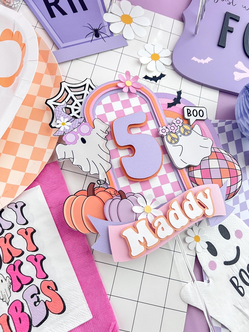 May include: A layered cake topper featuring the number 5 in lavender, a checkered arch, two ghosts with accessories, a disco ball, pumpkins, and a banner with the name Maddy. The design includes a spiderweb, small flowers, and bats in pastel colors.