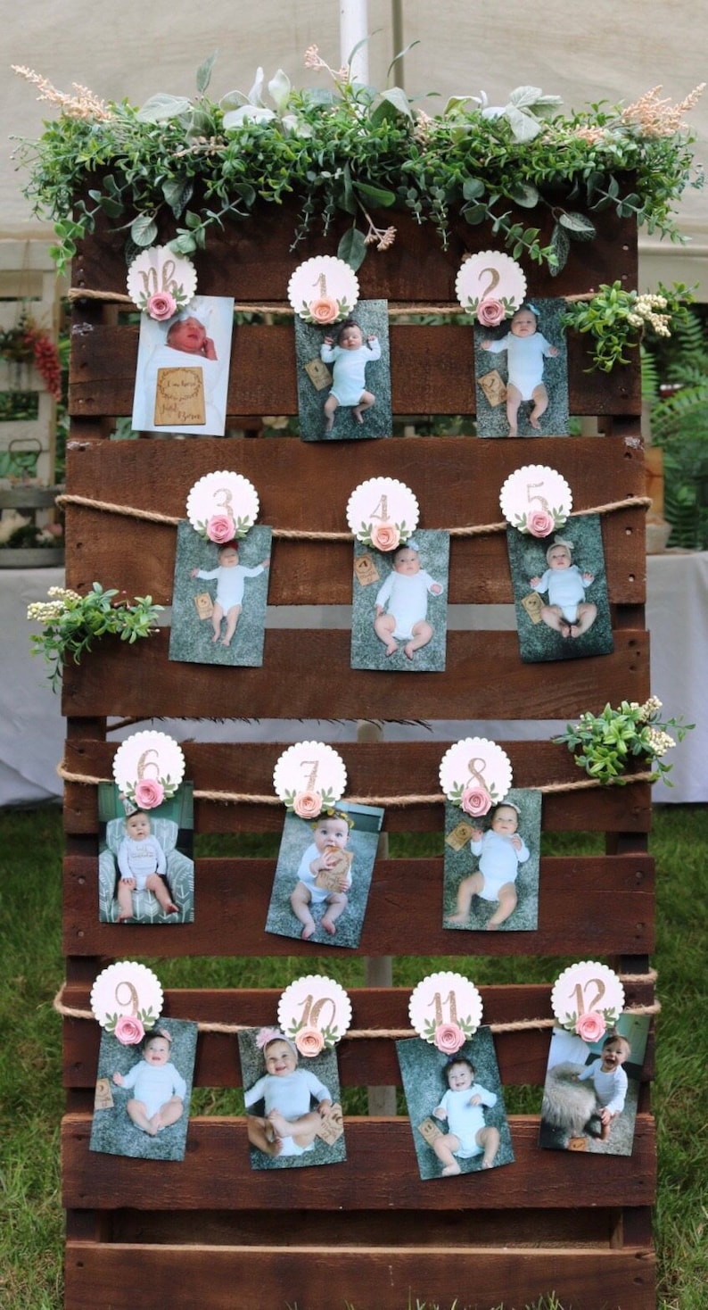 May include: A wooden pallet with twelve photos of a baby displayed in a grid pattern. Each photo is attached to a white circle with a pink flower and a gold number from 1 to 12. The photos are arranged in rows of three. The pallet is decorated with green foliage.