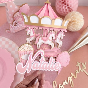 May include: A pink and white carousel cake topper featuring three horses, a popcorn bucket, and cotton candy. The name Natalie is printed in large white and pink script lettering across the front. The structure has gold accents.