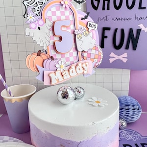 May include: A layered cake topper featuring a large number 5, a banner with the name Maddy, two ghosts, pumpkins, a disco ball, and a spiderweb. The design uses pastel colors including pink, purple, and orange with a checkered arch background.