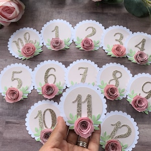 May include: Twelve round, pink and white table number cards with gold glitter numbers 1 through 12 and pink paper flowers. The table numbers are arranged in a semi-circle pattern.