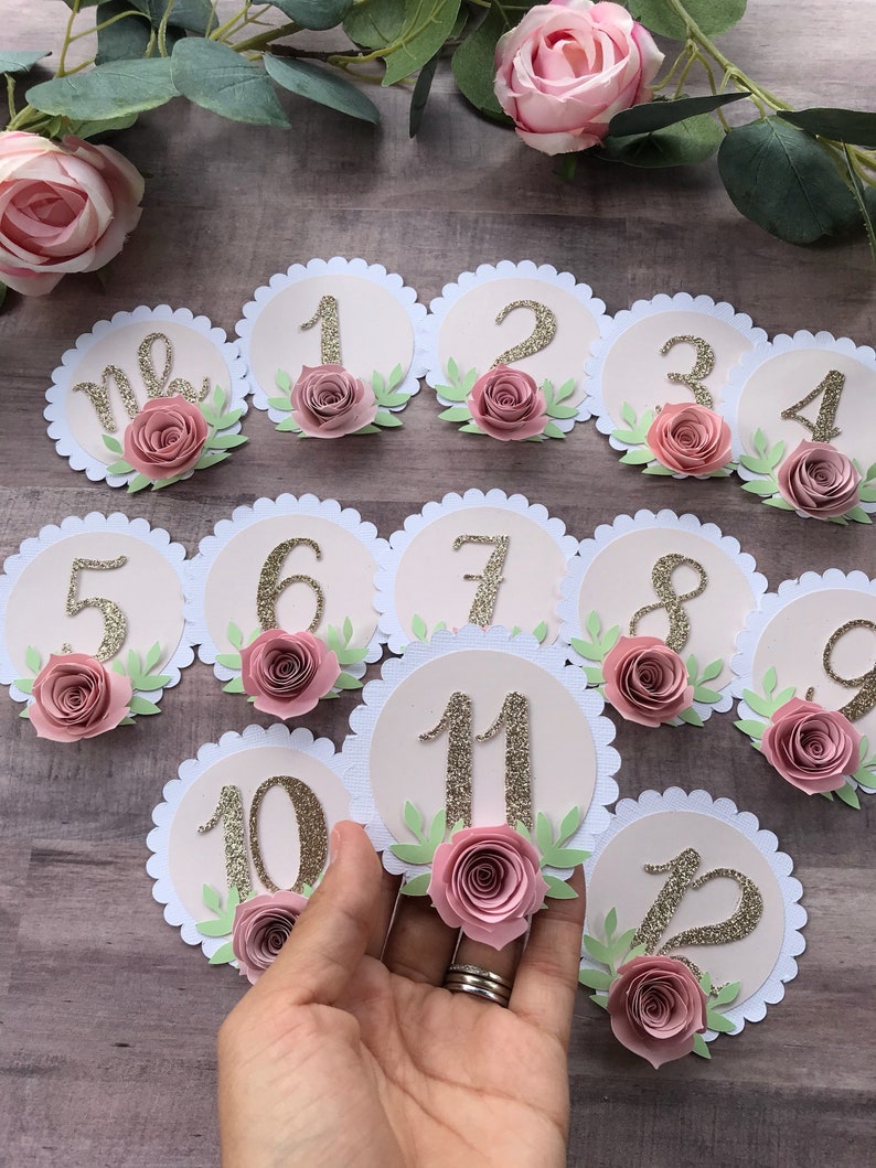 May include: Twelve round, pink and white table number cards with gold glitter numbers 1 through 12 and pink paper flowers. The table numbers are arranged in a semi-circle pattern.