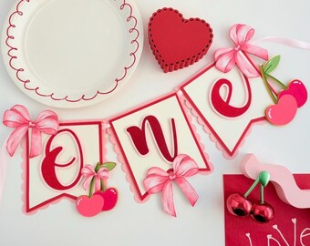 May include: A decorative banner spelling the word one with cream-colored pennants, red scalloped borders, pink ribbon bows, and cherry illustrations. Includes a white plate with a red scalloped rim, a red heart-shaped box, and red cherry ornaments.