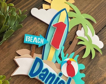 May include: A colorful beach-themed cake topper with a surfboard, palm trees, and a sun. The number "1" is displayed on the surfboard. The name "Daniel" is written on a banner. A beach ball and "Beach" sign are also included.