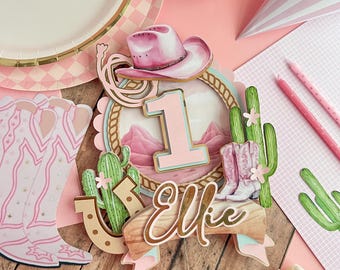 May include: Pink and gold cake topper with a cowboy hat, boots, cactus, horseshoe, and the number 1. The name "Ellie" is written in gold script on a wooden sign. The topper is on a wooden background with a pink "Happy Birthday" banner.