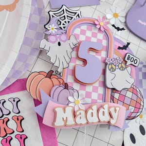 May include: A layered paper cake topper featuring the number 5, the name Maddy, two ghosts, pumpkins, a disco ball, and daisies. The design uses a pastel purple and orange checkered pattern with a spooky theme, including a spider web and bats.