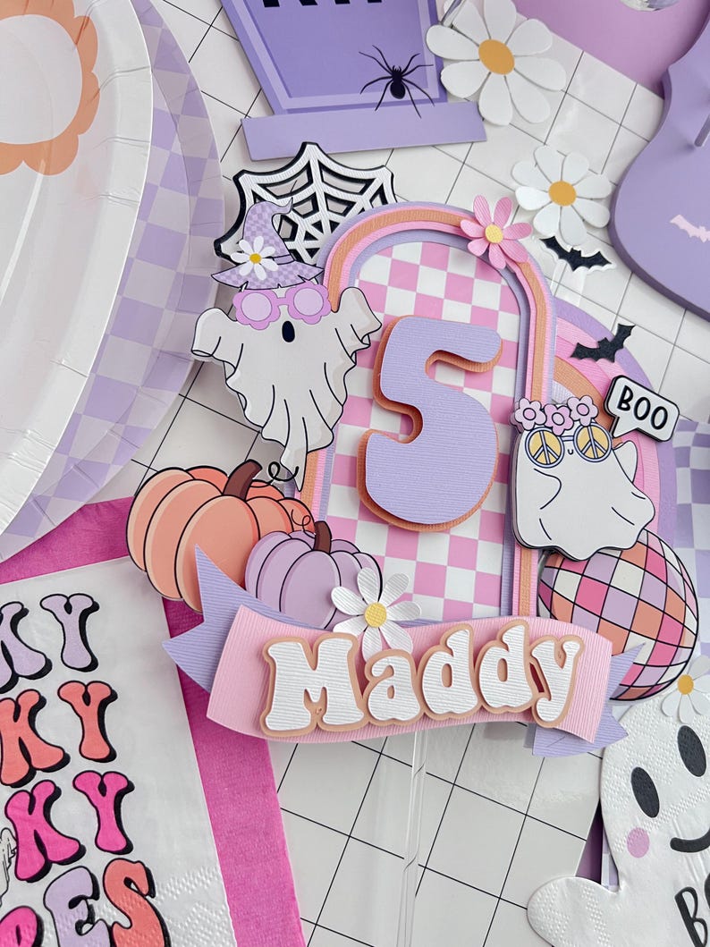 May include: A layered paper cake topper featuring the number 5, the name Maddy, two ghosts, pumpkins, a disco ball, and daisies. The design uses a pastel purple and orange checkered pattern with a spooky theme, including a spider web and bats.