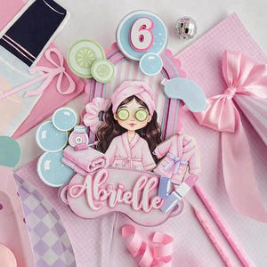 May include: A pink spa-themed cake topper featuring a cartoon girl in a robe with cucumber slices on her eyes. The topper includes the name "Abrielle", a number 6, and spa-related accessories. The background is a mix of pink and light blue elements.