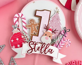 May include: Pink and white cake topper with a pink number one, a gingerbread man, a candy cane, a Christmas tree, a reindeer, a present, and a red and white ornament. The topper also includes a white banner with the name "Stella" in red.