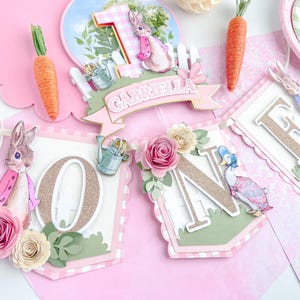 May include: A pink and green banner with the word "ONE" in gold glitter letters. The banner is decorated with flowers, a bunny, and a duck. The banner also includes the name "Gabriella".
