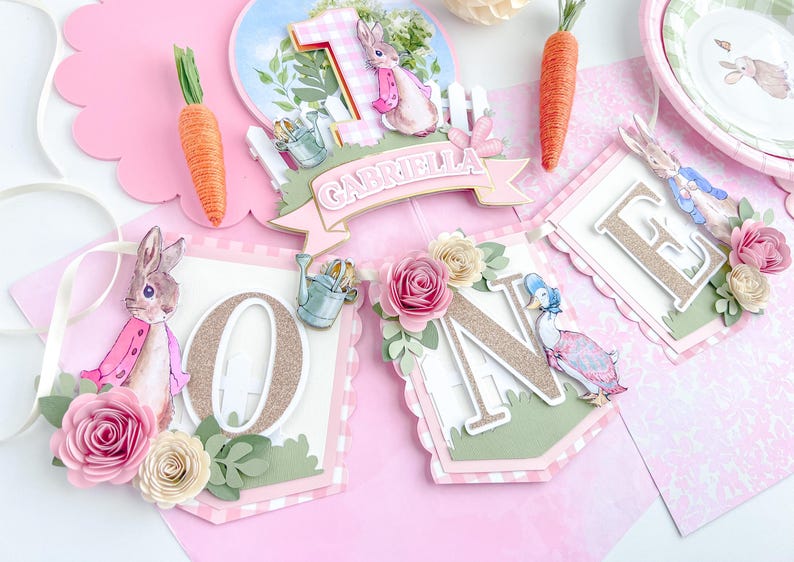May include: A pink and green banner with the word "ONE" in gold glitter letters. The banner is decorated with flowers, a bunny, and a duck. The banner also includes the name "Gabriella".
