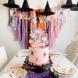 May include: A light pink cake on a black stand, decorated with plastic spiders and a custom topper featuring the number 7, the name Isabella, ghosts, a disco ball, and a spiderweb. The cake is surrounded by Halloween-themed party supplies.