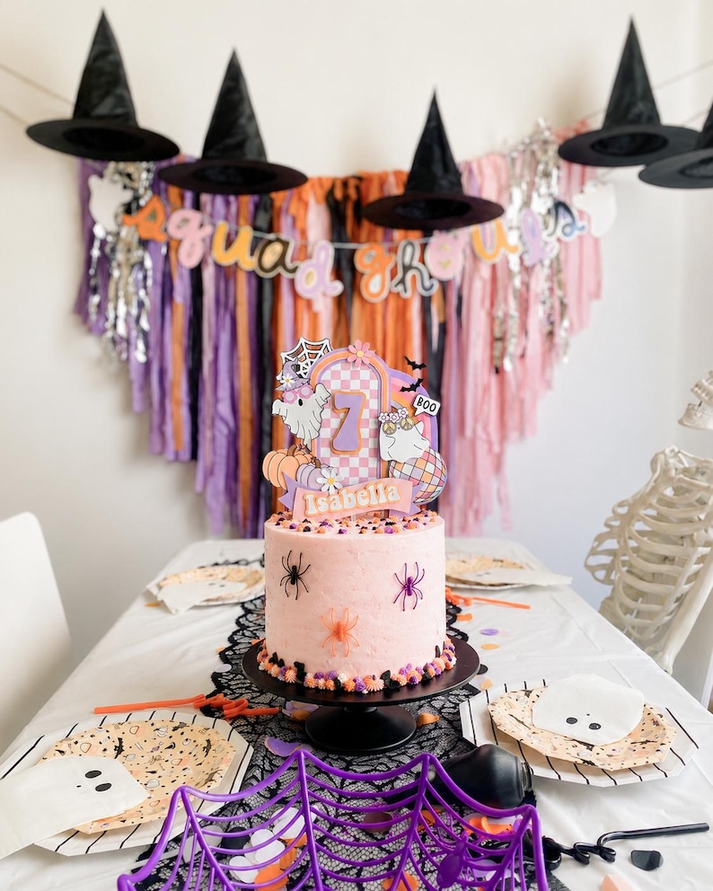 May include: A light pink cake on a black stand, decorated with plastic spiders and a custom topper featuring the number 7, the name Isabella, ghosts, a disco ball, and a spiderweb. The cake is surrounded by Halloween-themed party supplies.