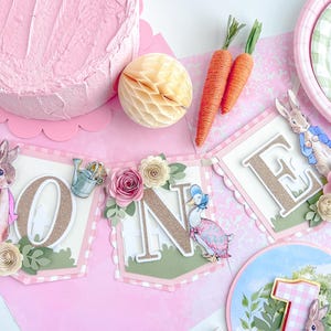 May include: A pink and green banner with the word "ONE" in gold glitter letters. The banner is decorated with flowers, leaves, and a rabbit and duck character.
