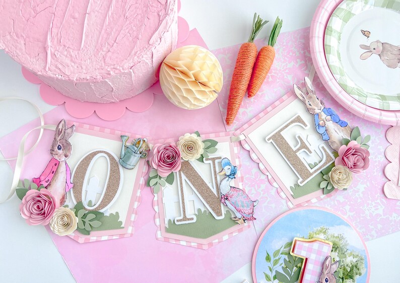 May include: A pink and green banner with the word "ONE" in gold glitter letters. The banner is decorated with flowers, leaves, and a rabbit and duck character.