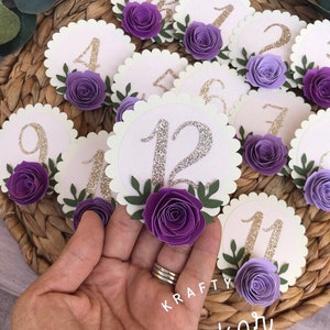 May include: Purple and white paper flower table number cards with gold glitter numbers. The numbers range from 1 to 12. The cards have scalloped edges and are decorated with green leaves.