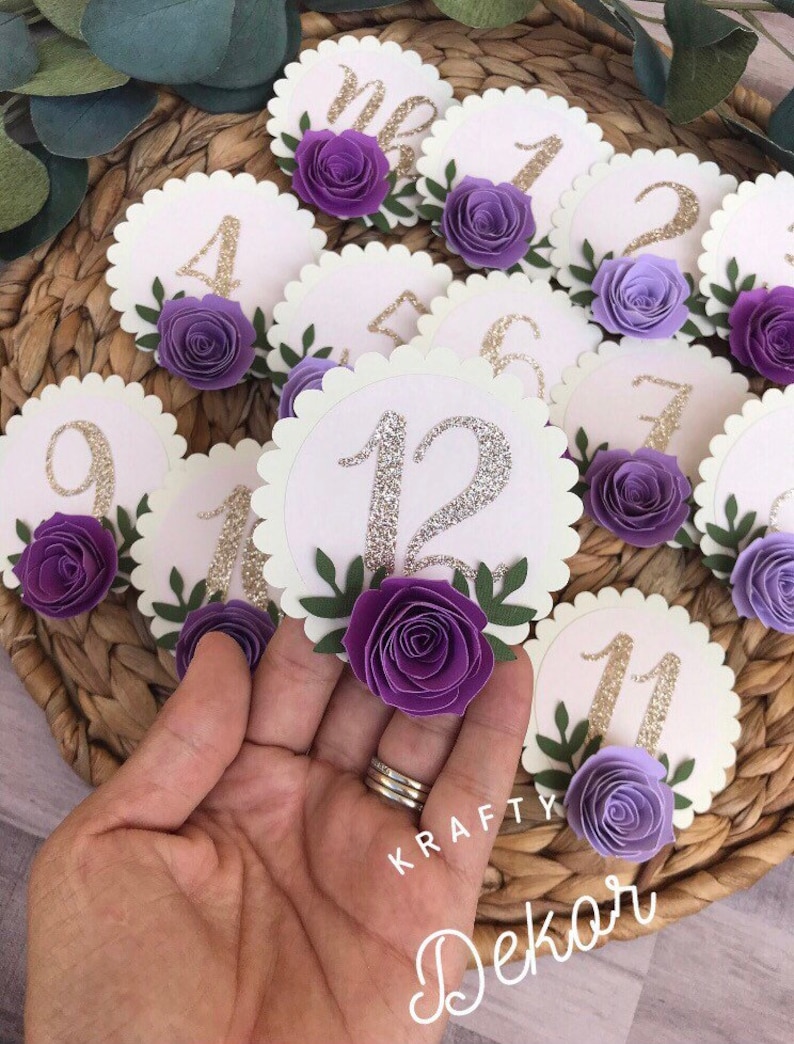 May include: Purple and white paper flower table number cards with gold glitter numbers. The numbers range from 1 to 12. The cards have scalloped edges and are decorated with green leaves.