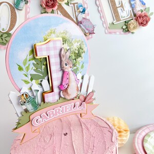 May include: A pink and white cake topper featuring a bunny rabbit with a pink dress and a pink and white gingham number one. The topper also includes a green watering can, a white picket fence, and a pink banner with the name "Gabriella".
