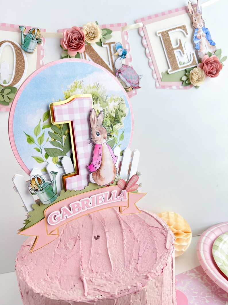 May include: A pink and white cake topper featuring a bunny rabbit with a pink dress and a pink and white gingham number one. The topper also includes a green watering can, a white picket fence, and a pink banner with the name "Gabriella".