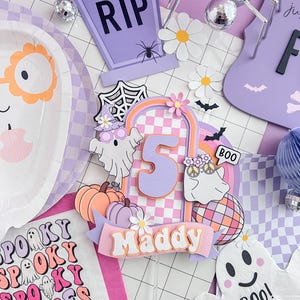 May include: A layered paper cake topper featuring the number 5 in orange, a checkered arch, two ghosts, pumpkins, a disco ball, and a banner with the name Maddy. The design includes pastel purple, orange, and white colors with floral accents.