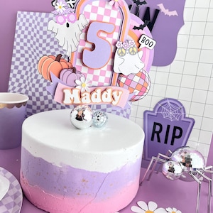 May include: A round cake with white, lavender, and pink ombre frosting, topped with a layered paper cake topper featuring the number 5, the name Maddy, ghosts, pumpkins, a disco ball, and flowers, accented by small silver disco ball decorations.