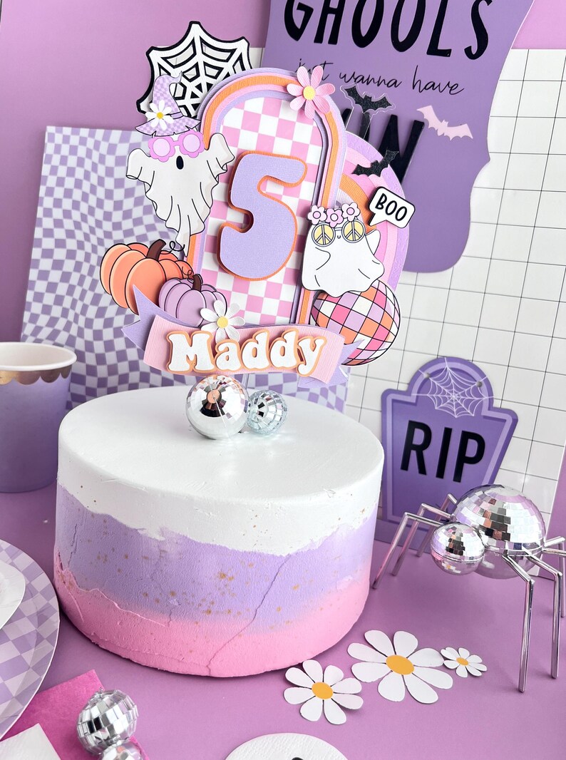 May include: A round cake with white, lavender, and pink ombre frosting, topped with a layered paper cake topper featuring the number 5, the name Maddy, ghosts, pumpkins, a disco ball, and flowers, accented by small silver disco ball decorations.