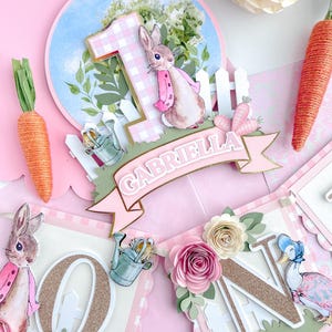 May include: A pink and green banner with the letters "ONE" in gold glitter. The banner features a bunny rabbit, a watering can, and flowers. The banner also includes the name "GABRIELLA" in gold glitter.