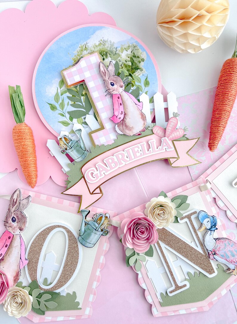 May include: A pink and green banner with the letters "ONE" in gold glitter. The banner features a bunny rabbit, a watering can, and flowers. The banner also includes the name "GABRIELLA" in gold glitter.