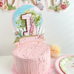 May include: A pink and white cake with a Peter Rabbit themed topper. The topper features a bunny, a number one, a fence, and a banner that says "Gabriella".