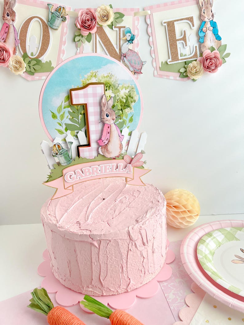 May include: A pink and white cake with a Peter Rabbit themed topper. The topper features a bunny, a number one, a fence, and a banner that says "Gabriella".