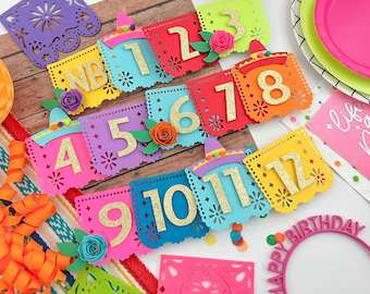 May include: A colorful paper banner with numbers 1 through 12, decorated with gold glitter and small paper flowers. The banner is perfect for a birthday party.