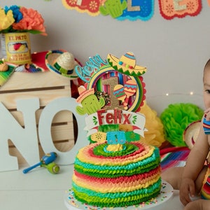 May include: A colorful birthday cake with a festive Mexican theme. The cake is topped with a decorative topper featuring a sombrero, cactus, and the name "Felix". A banner spelling "Feliz Cumpleaños" is in the background.