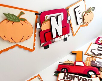 May include: A red truck with a pumpkin in the back, a white letter N, and the word "ONE" in orange letters. The truck is part of a banner with orange and white scalloped edges. There are pumpkins on each side of the truck.