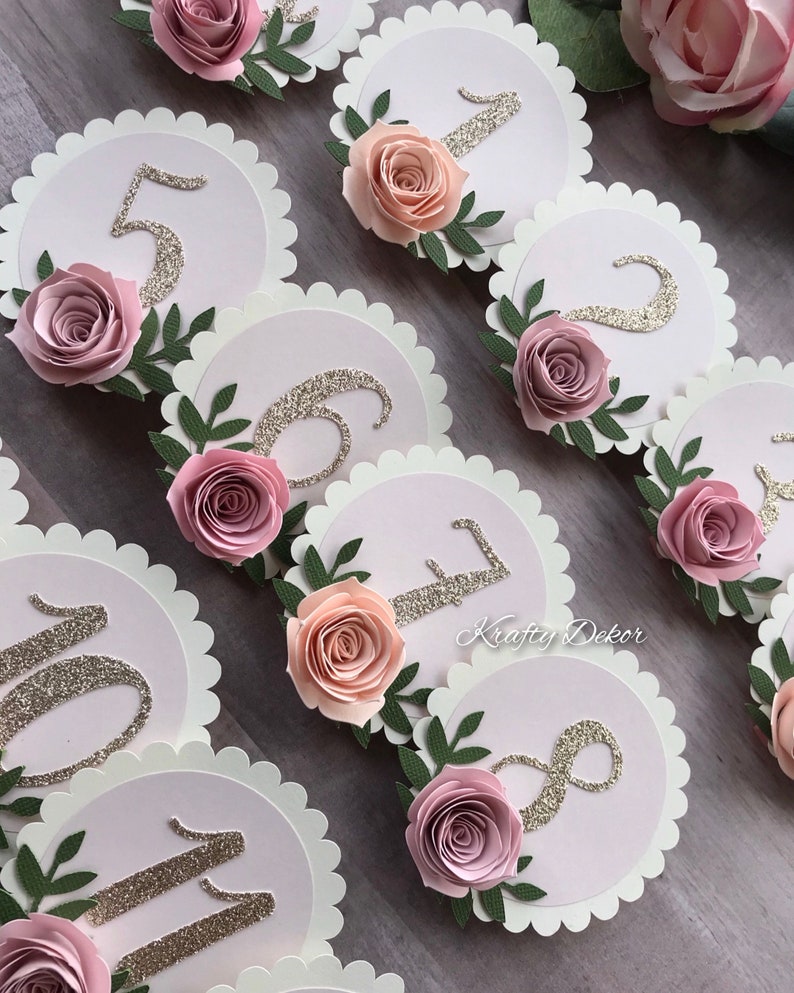 May include: Pink and white paper circles with gold glitter numbers 1 through 11. Each circle has a pink paper flower and green leaves. The text 'Krafty Dekor' is visible on one of the circles.